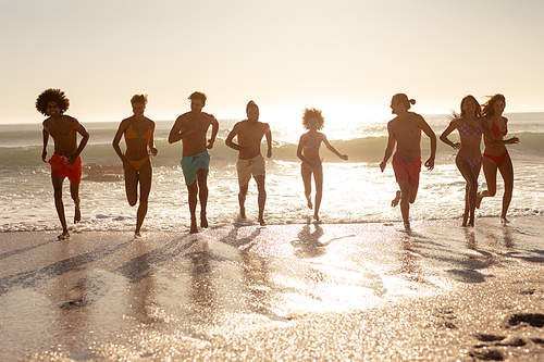 A multi-ethnic group of male and female friends on holiday having fun on a beach, running out of the sea as the sun goes down behind them