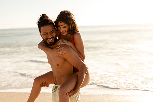 Portrait of a tanned Caucasian couple on holiday, having fun piggybacking on a sunny beach, looking to camera and smiling, with blue sky and sea in the background