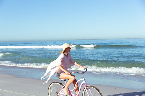 A Caucasian woman wearing a hat enjoying time at the beach on a sunny day, riding a bike, with sea in the background