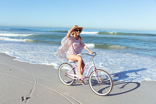 A Caucasian woman wearing a hat enjoying time at the beach on a sunny day, riding a bike, with sea in the background