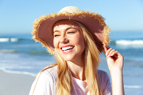 Close up of a Caucasian woman enjoying time at the beach on a sunny day, wearing a hat, smiling, with sea in the background