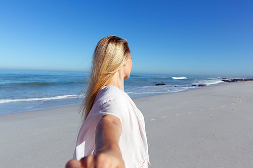 A Caucasian woman enjoying time at the beach on a sunny day, reaching to camera and looking away, with sea in the background