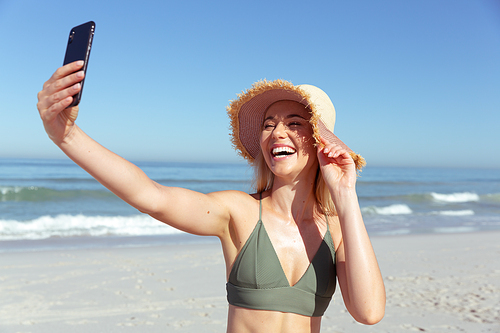 A Caucasian woman enjoying time at the beach on a sunny day, taking a selfie and smiling, with sea in the background