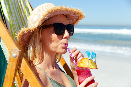 Close up of a Caucasian woman wearing a hat enjoying time at the beach on a sunny day,  sitting on deck chair, drinking cocktail