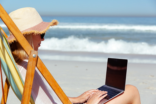 A Caucasian woman wearing a hat enjoying time at the beach on a sunny day,  sitting on deck chair, using a laptop computer