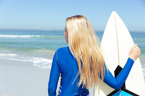 A Caucasian woman enjoying time at the beach on a sunny day, standing and holding a surfboard, with sea in the background