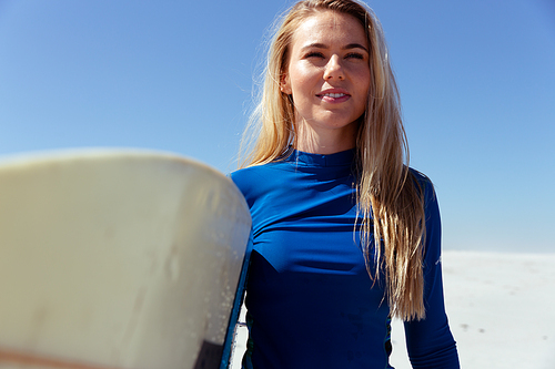 Close up of a Caucasian woman enjoying time at the beach on a sunny day, holding surfboard and walking, with blue sky in the background