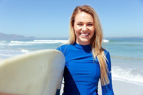 Portrait of a Caucasian woman enjoying time at the beach on a sunny day, holding surfboard and walking,  and smiling, with sea in the background