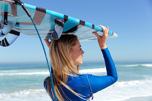 A Caucasian woman enjoying time at the beach on a sunny day, holding surfboard and walking, with sea in the background