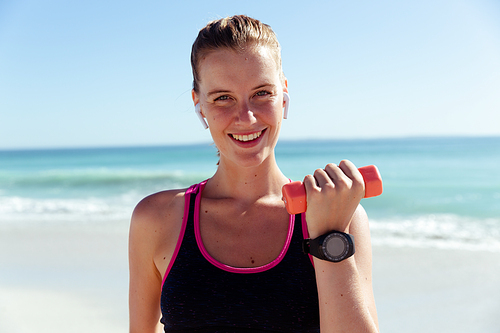Portrait of a Caucasian woman wearing sports clothes, enjoying time at the beach on a sunny day,  and smiling, holding dumbbells, with sea in the background