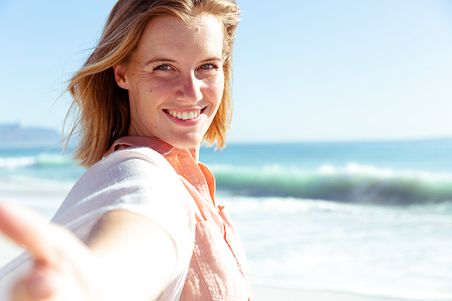 Portrait of a Caucasian woman enjoying time at the beach on a sunny day, reaching to camera and smiling, with sea in the background