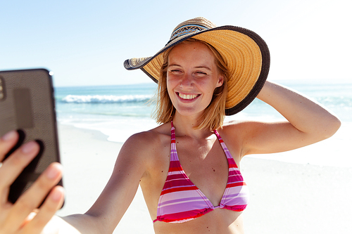 A Caucasian woman wearing hat, enjoying time at the beach on a sunny day, taking a selfie and smiling, with sea in the background
