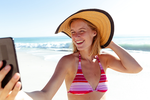 A Caucasian woman wearing hat, enjoying time at the beach on a sunny day, taking a selfie and smiling, with sea in the background