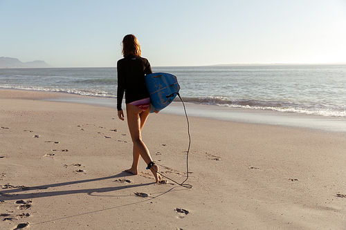 A Caucasian woman enjoying time at the beach on a sunny day, holding surfboard and walking, with sea in the background