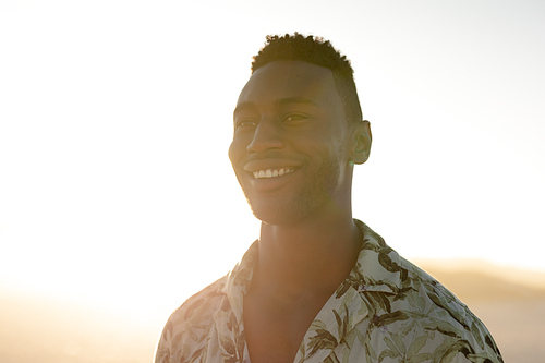 A happy African American man enjoying free time on beach on a sunny day, standing on sand with sun shining behind him, wearing a Hawaiian shirt. Relaxing summer vacation.