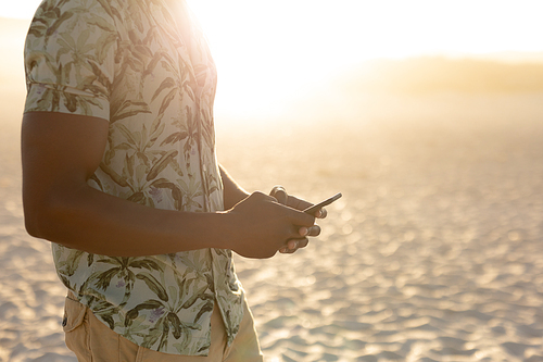 An African American man enjoying free time on beach on a sunny day, standing on sand with sun shining behind him, wearing a Hawaiian shirt, using his smartphone. Relaxing summer vacation.