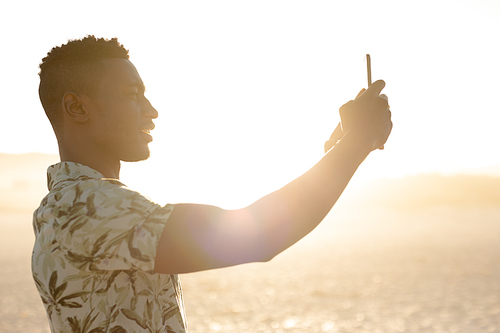 An African American man enjoying free time on beach, standing on sand with sun shining behind him, wearing a Hawaiian shirt, using his smartphone, taking photos. Relaxing summer vacation.