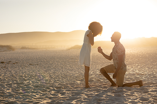 Mixed race couple enjoying free time on beach on a sunny day together, man proposing to the woman, kneeling on one knee on the sand. Relaxing summer vacation.