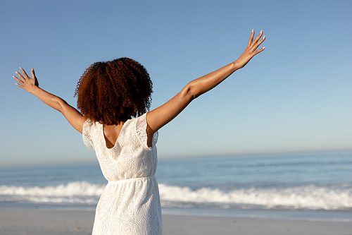 A rear view of a happy, attractive mixed race woman enjoying free time on beach on a sunny day, wearing a white dress, sun shining on her face with her arms outstretched. Relaxing summer vacation.