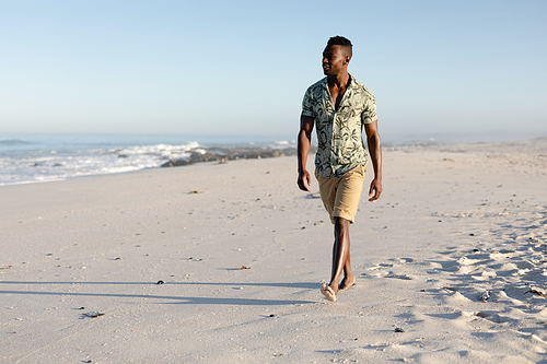 Attractive African American man enjoying free time on beach on a sunny day, wearing a Hawaiian shirt, walking on the beach sun shining on his face. Relaxing summer vacation.