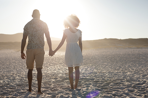 A rear view of a mixed race couple enjoying free time on beach on a sunny day together, holding hands with sun shining on their faces.