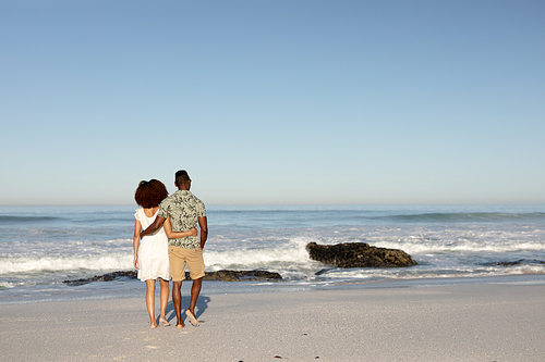 A rear view of a mixed race couple enjoying free time on beach on a sunny day together, walking and holding each other with sun shining behind them. Relaxing summer vacation.