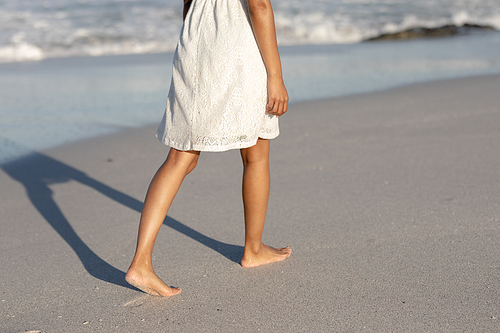 A happy, attractive mixed race woman enjoying free time on beach on a sunny day, wearing a white dress, walking, sun shining on her, bare feet touching the sand. Relaxing summer vacation.