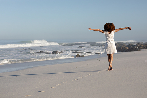 A rear view of a happy, attractive mixed race woman with her arms outstretched enjoying free time on beach, wearing a dress, facing the sea, sun shining on behind her.  Relaxing summer vacation.