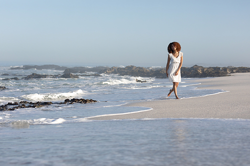A happy, attractive mixed race woman enjoying free time on beach on a sunny day, wearing a white dress, walking, sun shining on her face. Relaxing summer vacation.
