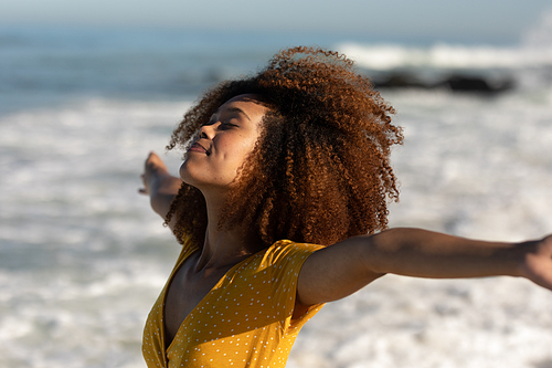 A rear view of a happy, attractive mixed race woman with her arms outstretched enjoying free time on beach, wearing a dress, facing the sea, sun shining on behind her.  Relaxing summer vacation.