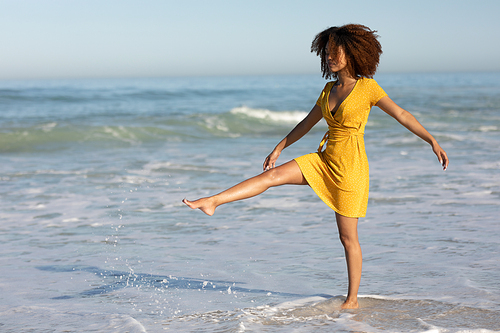 A happy, attractive mixed race woman enjoying free time on beach on a sunny day, wearing a yellow dress, playing with water with her leg in the air, sun shining on her face. Relaxing summer vacation.