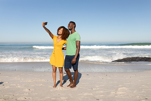 A mixed race couple enjoying free time on beach on a sunny day together, taking a selfie, smiling and posing with sun shining on their faces. Relaxing summer vacation.