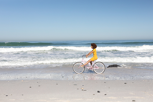 A happy, attractive mixed race woman enjoying free time on beach on a sunny day, wearing a yellow dress, riding a bicycle, sun shining on her face. Relaxing summer vacation.