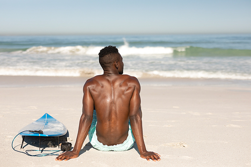 A rear view of an attractive African American man enjoying free time on beach on a sunny day, smiling, having fun, surfing, sitting on the beach, sun shining on his face.