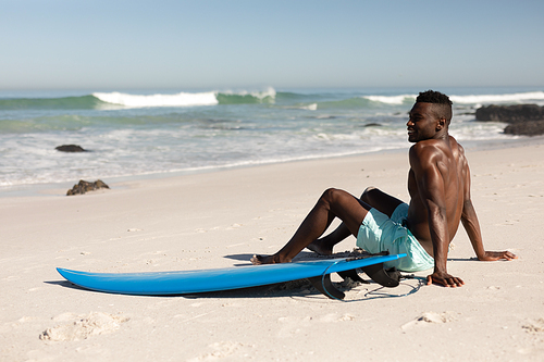 An attractive African American man enjoying free time on beach on a sunny day, smiling, having fun, surfing, sitting on the beach, sun shining on his face.