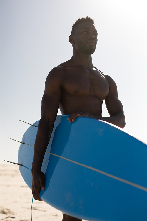An attractive African American man enjoying free time on beach on a sunny day, having fun, surfing, holding a surfboard, sun shining behind him.
