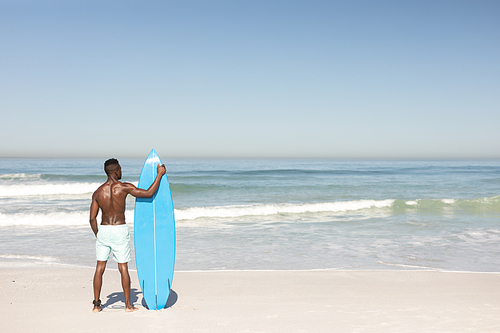 A rear view of an attractive African American man enjoying free time on beach on a sunny day, smiling, having fun, standing with his surfboard, sun shining on him.