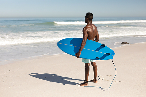 An attractive African American man enjoying free time on beach on a sunny day, smiling, having fun, standing with his surfboard, sun shining on him.