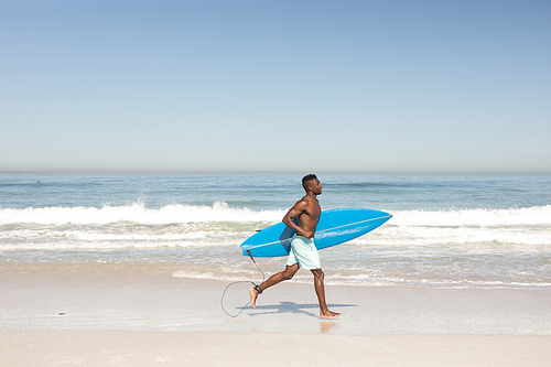 An attractive African American man enjoying free time on beach on a sunny day, smiling, having fun, running with his surfboard, sun shining on him.