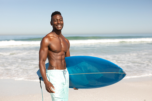 A portrait of a happy, attractive African American man enjoying free time on beach on a sunny day, smiling, having fun, standing with his surfboard, sun shining on him.