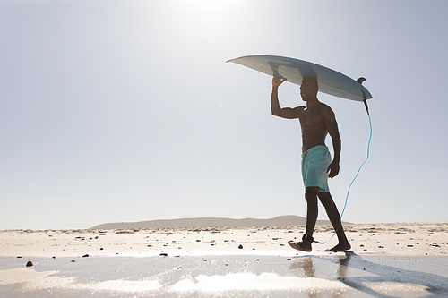A happy, attractive African American man enjoying free time on beach on a sunny day, having fun, carrying his surfboard, sun shining on him.