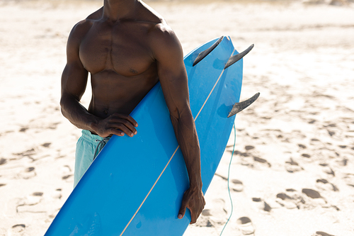 An attractive African American man enjoying free time on beach on a sunny day, having fun, carrying his surfboard, sun shining on him.