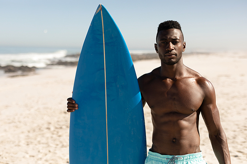 A portrait of an attractive African American man enjoying free time on beach on a sunny day, smiling, having fun, standing with his surfboard, sun shining on him.