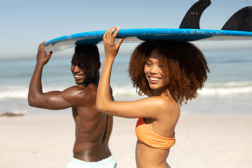 A portrait of a mixed race couple enjoying free time on beach on a sunny day together, surfing, carrying their surfboards, having fun with sun shining on their faces.