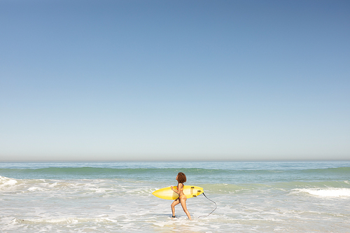 an attractive mixed race woman enjoying free time on beach on a sunny day, wearing a swimsuit, walking on the sand, surfing, carrying her surfboard, sun shining on her face.