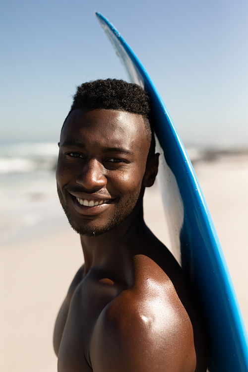 A portrait of a happy, attractive African American man enjoying free time on beach on a sunny day, having fun, standing with his surfboard, sun shining on him.