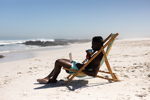 An attractive African American man enjoying free time on beach on a sunny day, drinking, sitting in a deck chair, using his laptop.