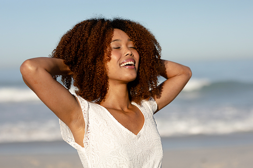A mixed race woman with her arms behind her head enjoying free time on beach on a sunny day, wearing white dress, sun shining on her face.