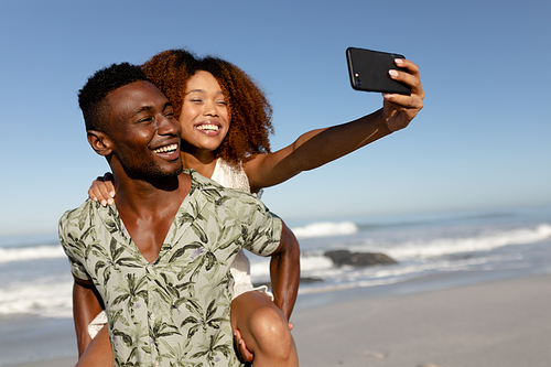A mixed race couple enjoying free time on beach on a sunny day together, piggy backing, laughing and taking photos with a smartphone with sun shining on their faces.