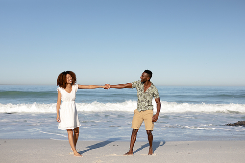 A mixed race couple enjoying free time on beach on a sunny day together, walking and holding each other with sun shining on their faces.